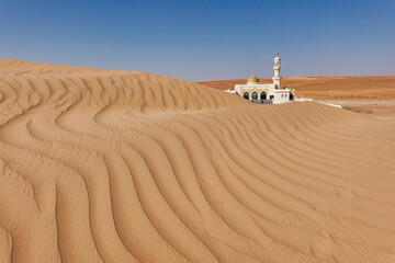 In the background is a white mosque semi-covered by a Wahiba desert dune in the foreground waves of...