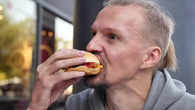 A handsome young Caucasian man bites off a juicy cheeseburger with a voracious appetite, satisfying his hunger during his lunch break at work, using fast food delivery.