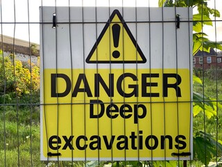 Yellow and white construction sign "Danger Deep Excavations" attached to a metal fence