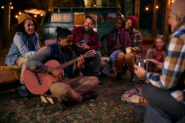 Happy black man playing acoustic guitar while camping with family and friends in forest.