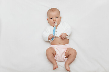 Infant baby girl in a white knitted blouse and pink knitted underpants is lying on back on a white blanket and playing with teething toy. Close up. 