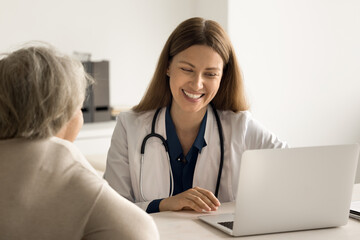 Smiling young doctor in uniform sit at desk talk to female retied client at medical checkup, show test result, explain treatment disease. Caring therapist use laptop to review patient health history - obrazy, fototapety, plakaty