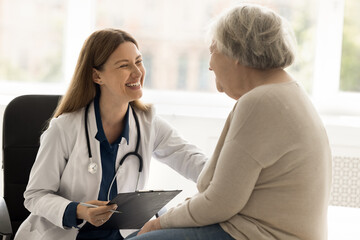 Caring young female therapist in white uniform gives support older woman, take care of patient, listen to health complaint. Retired patient enjoy good medical care and friendly attitude. Geriatrics