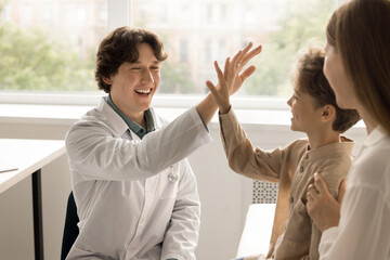 Male professional medical worker in white coat gives high five to child. Little kid with mother celebrates successful treatment, good medical result at check up meeting in clinic. Pediatry, childcare