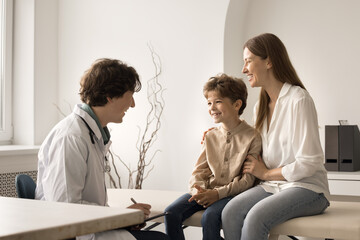 Little cute boy visit pediatrician doctor with loving mom. Friendly male paediatrician in coat hold clipboard writes symptoms, listen to kid patient, provide consultation. Healthcare, medical check-up