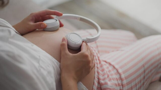 A pregnant girl in maternity clothes is full of love trying to bring music to the unborn child through headphones in the room. Caucasian girl future newborn listens to music through headphones