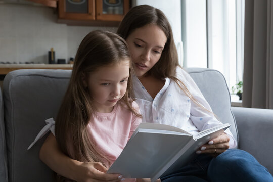 Pretty loving mom hugging serious little kid girl reading book to daughter on couch, teaching to read. Bored kid listening to fairy tale mother telling. Motherhood, preschool education concept