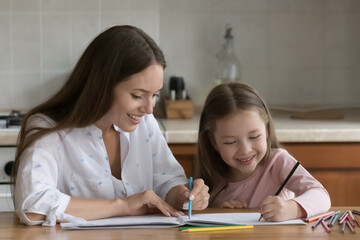 Happy daycare teacher teaching little kid girl to draw, sitting at home kitchen table. Mom and little daughter making doodles in paper album, scratching with colorful pencils, talking, laughing