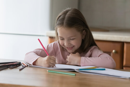 Happy cute little kid girl drawing in colored pencils, sitting at kitchen table, scratching doodles in paper album, training creative artistic skills. Child enjoying home activities