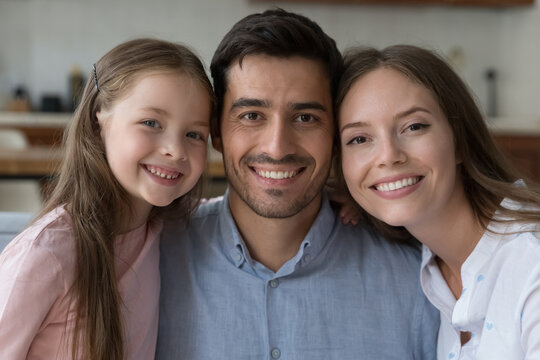 Happy attractive couple of parents and pretty little daughter kid head shot portrait. Young mother, father, child girl looking at camera with toothy smiles, sitting close, hugging. Family concept