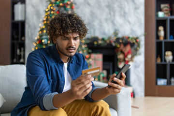 Cheated and upset Christmas man in living room near decorated Christmas tree, rejected and wrong money transfer, hispanic man holding bank credit card and phone. - obrazy, fototapety, plakaty