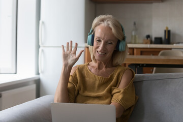 Happy senior 50s woman wearing wireless headphones using laptop computer, talking to family on video call. Mature blogger lady waving hand hello at webcam, greeting blog audience © fizkes