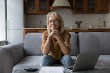Cheerful happy middle aged mature landlady woman doing monthly accounting paperwork, sitting on...