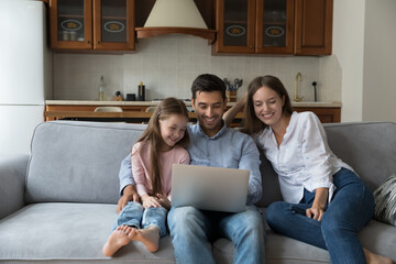 Cheerful laughing family couple of parents and little kid using online app on laptop, resting on...
