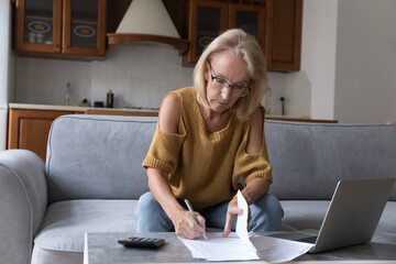 Focused serious middle aged homeowner in glasses, reviewing domestic documents, bills for payment, writing notes in papers at calculator and laptop, counting savings, taxes, insurance fees - obrazy, fototapety, plakaty