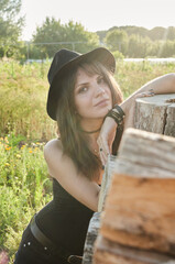 A girl in black clothes and a hat leans on a mountain of firewood, she is happy, and smiling, it&rsquo;s sunny outside, the weather is good. Relaxing at home in the garden.