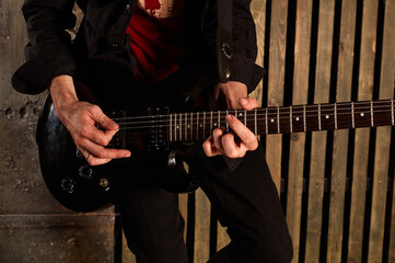 A male musician dressed in black plays a guitar against the background of a wooden wall. Hands holding a musical instrument. Performance.