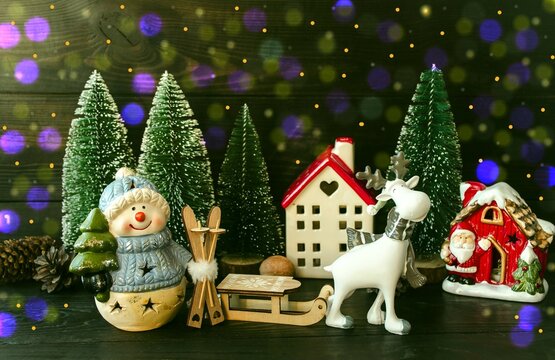 New Year's card, porcelain toy snowman with a Christmas tree in his hands and forest houses, white deer, green Christmas trees and wooden sleigh.  Home decoration.  Background picture.