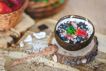 White yogurt with strawberries, blueberries, mint, grated coconut and chocolate in a coconut bowl on the kitchen table.