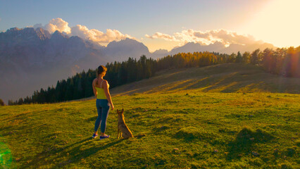 AERIAL: Attractive woman admires view of the mountains on a hilltop with her dog - obrazy, fototapety, plakaty