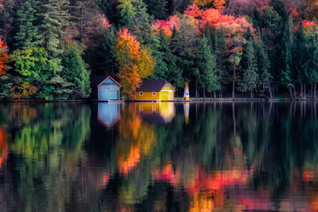 Autumn colors reflect in the waters at Old Forge Pond in NY.  A Boat House, shed and lighthouse on the opposite shore look beautiful in Autumn Colors. - obrazy, fototapety, plakaty