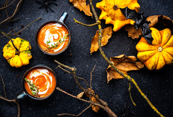Obraz na płótnie Canvas Festive creamy pumpkin soup with and .thyme. Autumn vegetarian food. Soup mug on black background with fallen oak leaves, pumpkins, spiders, twisted branches and bats. Top view