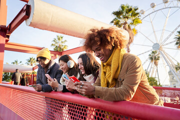 Smiling young persons using cell phones. Teenagers addicted to online technological trends. Group of friends leaning on a railing checking their social networks. People gathered at the amusement park. - obrazy, fototapety, plakaty