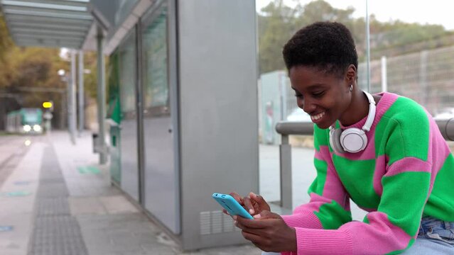 Happy young trendy african woman using mobile phone while waiting for the train at outdoors station. City life style and commuter people concept