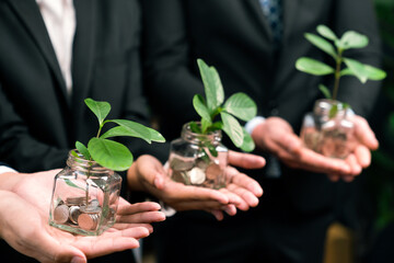 Business people holding money savings jar filled with coins and growing plant for sustainable...