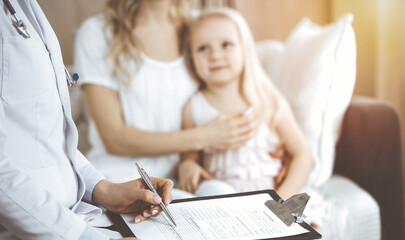 Doctor and patient. Pediatrician using clipboard while examining little girl with her mother at home. Happy cute caucasian child at medical exam. Medicine concept