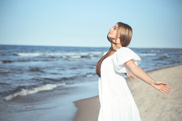 Fototapeta na wymiar Happy, beautiful woman on the ocean beach standing in a white summer dress, open arms