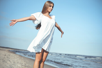 Happy, beautiful woman on the ocean beach standing in a white summer dress, open arms