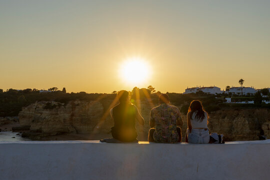 Young friends sitting on the wall, enjoying the sunset on a beach of Algarve, Portugal.