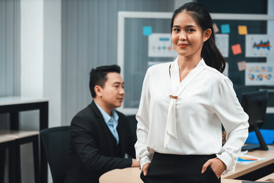 Successful businesswoman in white uniform standing in modern office in confident poses while smiling. Portrait of asian woman with arms akimbo. her partner working on laptop computer Intellectual.