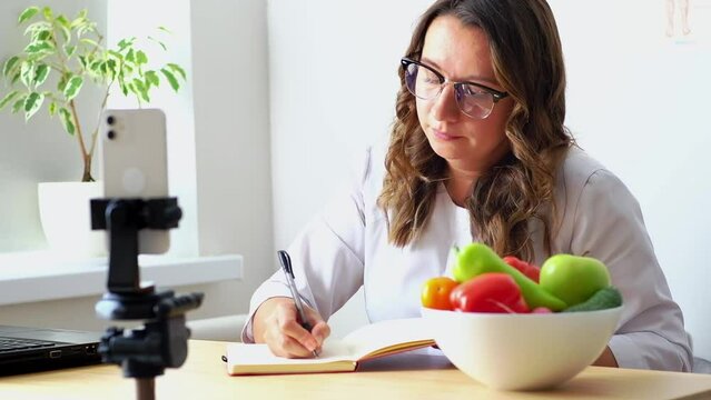 A female nutritionist sits at her desk and consults a patient over the phone and draws up a nutrition plan