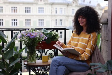 Young woman reading book at table on balcony with beautiful houseplants