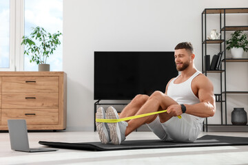 Fototapeta na wymiar Muscular man doing exercise with elastic resistance band near laptop on mat at home