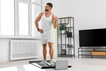 Muscular man doing exercise with elastic resistance band near laptop on mat at home