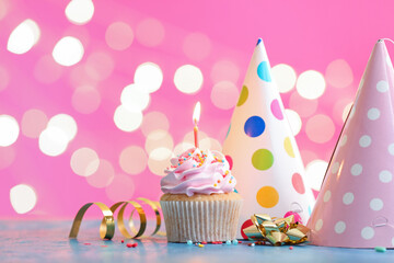 Tasty Birthday cupcake with burning candle and party hats on table against blurred lights