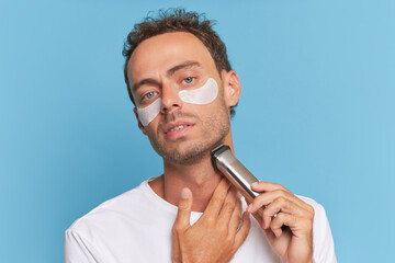 Young man does morning procedures, uses patches under eyes, shaves with electric razor, prepares for working day, male skincare concept, copy space