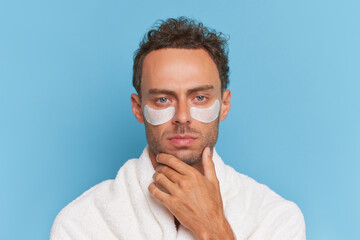 Close-up shot of guy uses cosmetics patches under eyes posing on blue background, cosmetic procedures for facial skin, handsome man concept, copy space