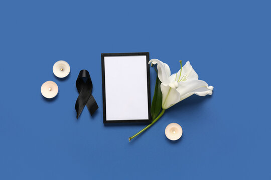 Black ribbon with white lily flower, candles and photo frame on blue background