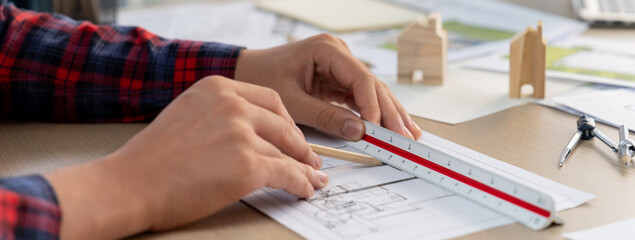 Closeup of architect engineer hand using ruler to mature and draw a blueprint on meeting table with wooden block, pencil and blueprint scatter around at architectural modern office. Delineation.