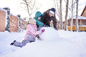 Fototapeta na wymiar A family builds a snowman out of white snow in the yard in winter.