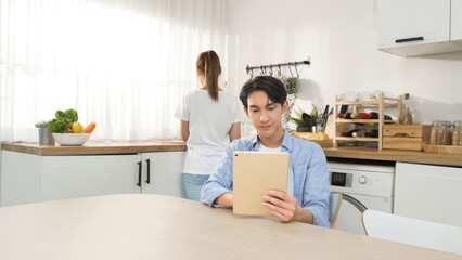 Obraz premium Asian home worker man focus on check email and to do list on tablet device while waiting young wife prepare healthy breakfast on a kitchen corner in the morning at home