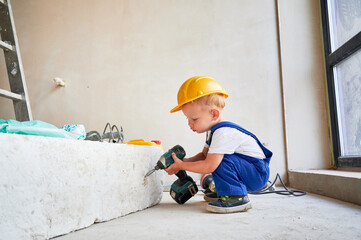 Adorable child in work overalls using electric construction tool in apartment under renovation. Cute kid in safety helmet working with power drill at home during renovation.