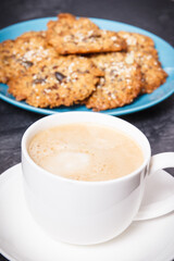 Cup of coffee with milk and fresh baked oatmeal cookies with honey and healthy seeds on blue plate. Delicious crunchy dessert