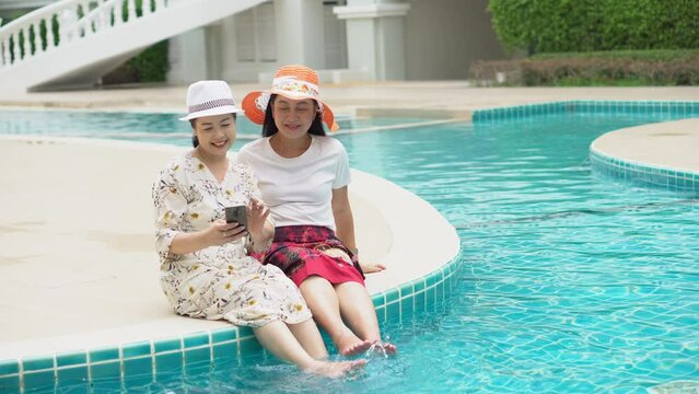 two senior asian woman with hat sitting by swimming pool using smart phone together outdoor. old lady take a photo . retired relaxation and vacation . friendship