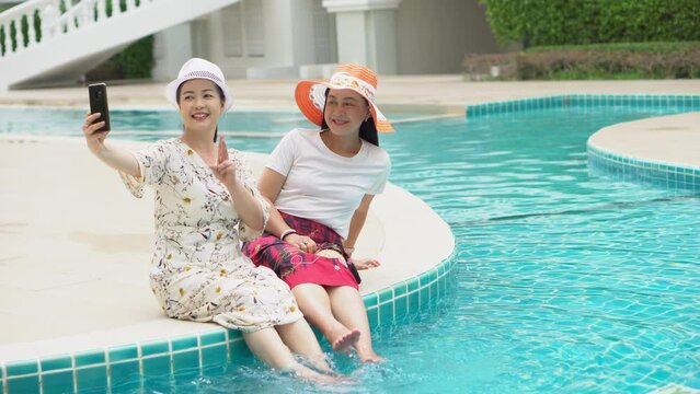 two senior asian woman with hat sitting by swimming pool take selfie with phone together outdoor. old lady take a photo . retired relaxation and vacation . friendship