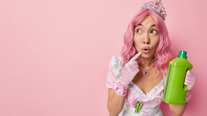 Obraz premium Studio shot of impressed female housemaid looks shocked aside keeps mouth opened holds green bottle of cleaning detergent wears crown dress and gloves isolated over pink background copy space on left